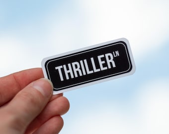 May include: A rectangular sticker with rounded corners, featuring the text "THRILLER" in white capital letters on a black background. The sticker is held against a blurred blue sky.