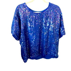 Puede incluir: Una blusa de manga corta con cuello redondo, cubierta de lentejuelas azules iridiscentes. Las lentejuelas capturan la luz, creando un efecto brillante. La blusa está colgada en una percha beige sobre un fondo blanco.