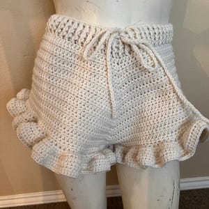 May include: Cream-colored crochet shorts with a drawstring waist and ruffled hem. The shorts are handmade with a textured stitch and a delicate, feminine design. The shorts are displayed on a mannequin.