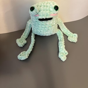 May include: A handmade, light green crochet frog with black eyes and pink cheeks. The frog has outstretched arms and legs, and a wide, black mouth. The plush toy is made of soft, textured yarn.