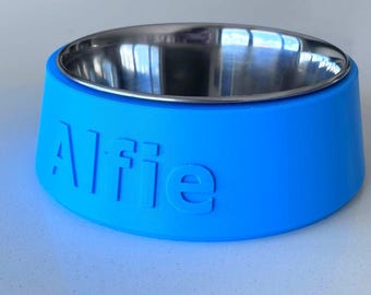 May include: A bright blue pet bowl with a stainless steel interior. The exterior of the bowl features the name Alfie embossed in large, raised letters on the side. The bowl has a wide, tapered base and a smooth, matte finish on the blue surface.