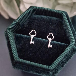Puede incluir: Un par de pendientes de botón plateados con forma de llave, presentados en una caja de joyería de terciopelo verde oscuro. Los pendientes son pequeños y delicados, con un diseño de llave clásico. La caja tiene forma hexagonal.