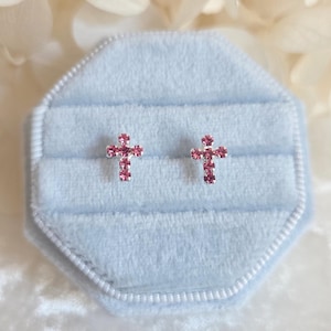 Puede incluir: Un par de pendientes de botón con forma de cruz, cada uno con pequeños cristales rosas brillantes engastados en una montura de metal plateado. Los pendientes se muestran uno al lado del otro, destacando el diseño de cruz y el color rosa.