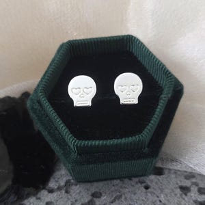 Op de afbeelding: Een paar zilveren schedeloorbellen met hartvormige ogen, gepresenteerd in een donkergroene fluwelen zeshoekige doos. De oorbellen zijn klein en gedetailleerd. Een uniek sieraad.