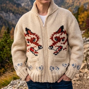 Könnte beinhalten: Cremefarbener Reißverschluss-Pullover mit Schalkragen und zwei Vordertaschen. Der Pullover hat ein rot-schwarzes Fuchsmotiv auf der Vorderseite und blaue Pfotenabdrücke am unteren Rand. Der Hintergrund zeigt eine Bergkette.