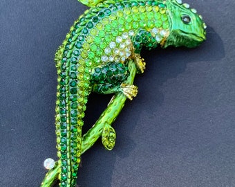 Könnte beinhalten: Eine grüne, mit Strass besetzte Chamäleon-Brosche mit klaren Strasssteinen, die auf einem Zweig mit grünen Blättern sitzt.