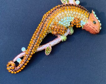 Könnte beinhalten: Eine goldfarbene Chamäleon-Brosche mit orangefarbenen und grünen Strasssteinen, die auf einem Zweig mit grünen Blättern sitzt.