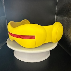 Peut inclure: Un casque jaune de style futuriste avec un détail en maille rouge. Il repose sur un support circulaire blanc sur fond sombre. Le design présente des formes arrondies et une forme symétrique.