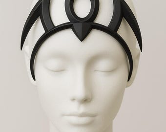May include: A black, geometric crown or headpiece with an open circle and curved accents. The headpiece is displayed on a white mannequin head. The design is symmetrical and appears to be made of a solid material.