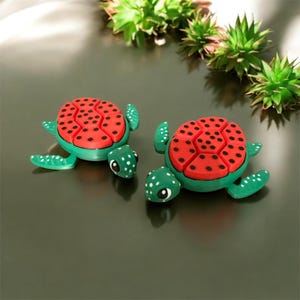 May include: Two green turtle figurines with red and black patterned shells. The turtles have white polka dots on their heads, flippers, and shells. The figurines are displayed on a dark surface with green plants in the background.