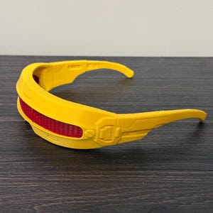 Può includere: Occhiali gialli in stile futuristico con una lente rossa a rete. Gli occhiali hanno un design curvo con aste allungate e un'estetica meccanica dettagliata. Gli occhiali sono probabilmente un accessorio di costume o un oggetto di scena.