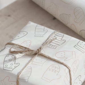 May include: A wrapped gift with a light-colored paper featuring a pattern of mittens in gray and beige. The gift is tied with twine. A roll of matching wrapping paper is in the background.