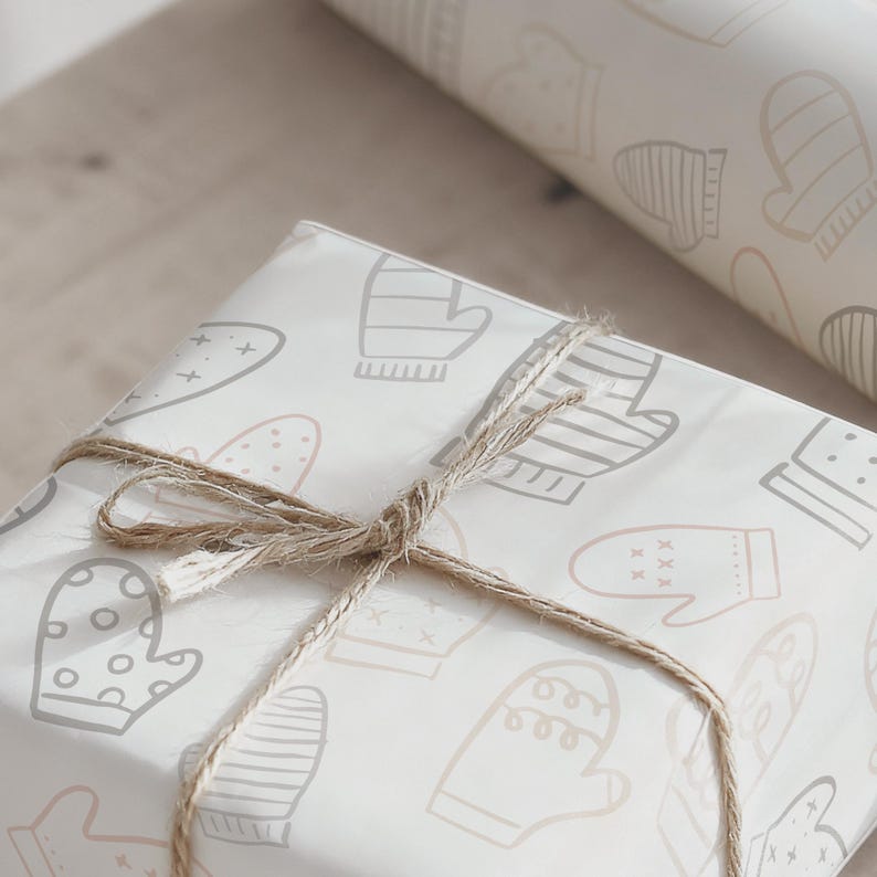 May include: A wrapped gift with a light-colored paper featuring a pattern of mittens in gray and beige. The gift is tied with twine. A roll of matching wrapping paper is in the background.