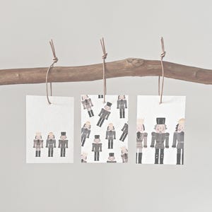 May include: Three rectangular gift tags with illustrations of nutcrackers hanging from a wooden branch. The tags feature different arrangements of the nutcracker figures, with varying poses and patterns. The tags are tied with brown twine.