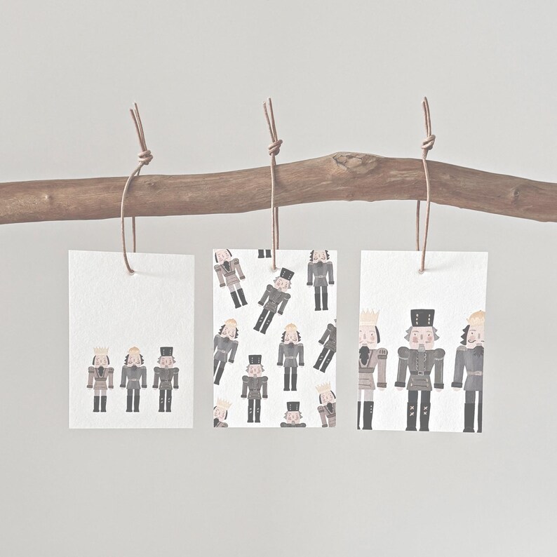May include: Three rectangular gift tags with illustrations of nutcrackers hanging from a wooden branch. The tags feature different arrangements of the nutcracker figures, with varying poses and patterns. The tags are tied with brown twine.