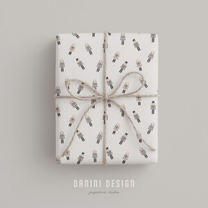 May include: A wrapped gift box with white paper featuring a repeating pattern of figures in coats and hats. The box is tied with twine in a bow. The text "DANINI DESIGN papeterie studio" is at the bottom.