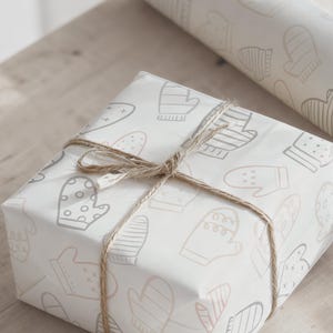 May include: A wrapped gift box with a light-colored paper featuring a pattern of mittens in gray and pink. The box is tied with twine. A roll of matching wrapping paper is visible in the background.