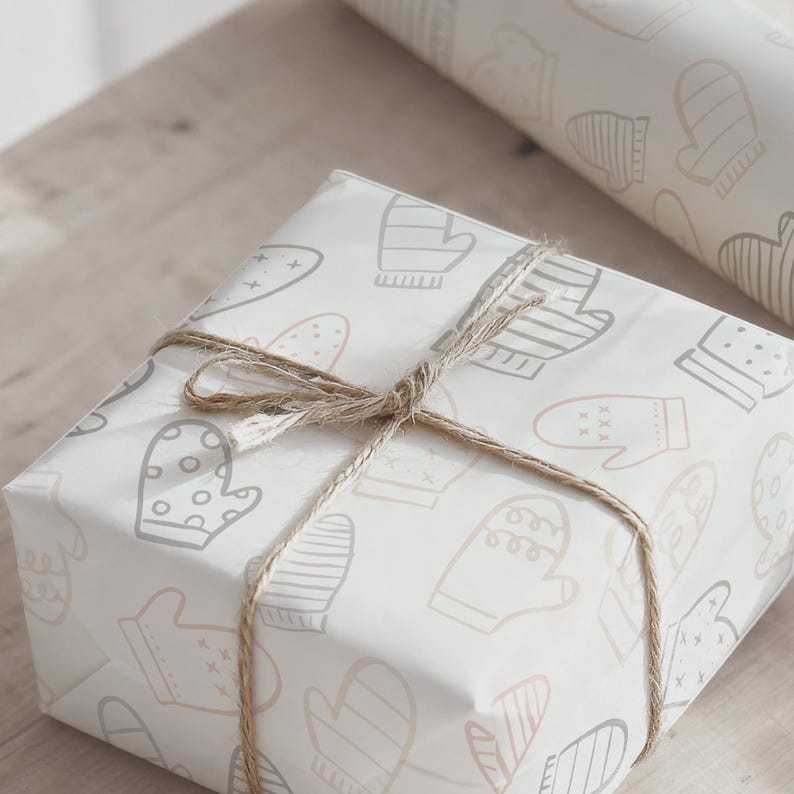 May include: A wrapped gift box with a light-colored paper featuring a pattern of mittens in gray and pink. The box is tied with twine. A roll of matching wrapping paper is visible in the background.