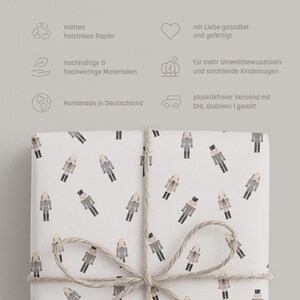 May include: White wrapping paper with a repeating pattern of nutcracker figures in gray, black, and tan. The paper is tied with twine. Text in German is visible in the background.