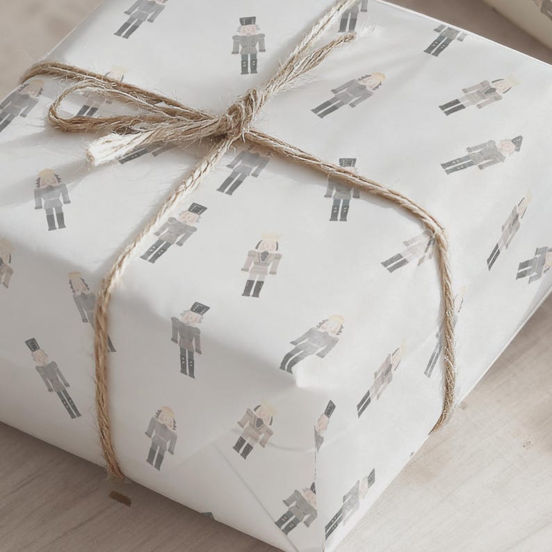 May include: A wrapped gift box featuring white paper with a repeating pattern of gray and beige nutcracker figures. The box is tied with natural twine, creating a rustic and festive presentation.