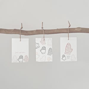 May include: Three white rectangular gift tags with mitten illustrations hang from a wooden branch. One tag has the text "Frohe Weihnachten" and mitten graphics. The other two tags have mitten patterns and the text "frohes fest".