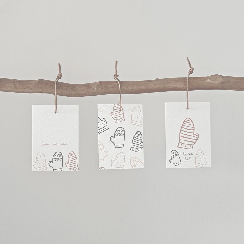 May include: Three white rectangular gift tags with mitten illustrations hang from a wooden branch. One tag has the text "Frohe Weihnachten" and mitten graphics. The other two tags have mitten patterns and the text "frohes fest".