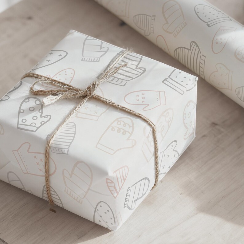 May include: A wrapped gift box with a light beige background and a pattern of mittens in various shades of gray and pink. The box is tied with twine. A roll of matching wrapping paper is in the background.
