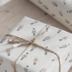 May include: A wrapped gift with a light-colored paper featuring a repeating pattern of gray nutcracker figures. The gift is tied with twine, creating a rustic and festive look. A roll of matching wrapping paper is visible in the background.