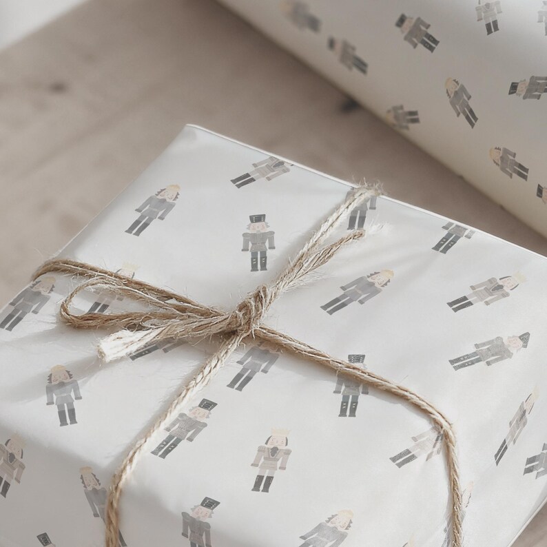 May include: A wrapped gift with a light-colored paper featuring a repeating pattern of gray nutcracker figures. The gift is tied with twine, creating a rustic and festive look. A roll of matching wrapping paper is visible in the background.