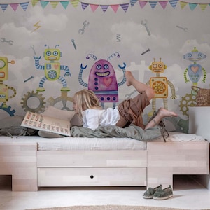 Op de afbeelding: Een kind leest op een licht houten bed met een grijze matras. De muur achter het bed heeft een grillige muurschildering van kleurrijke robots, tandwielen en gereedschappen tegen een bewolkte achtergrond. Een decoratieve slinger hangt bovenaan de muur.