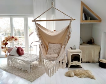 May include: A white macrame hammock chair with fringe hanging from the ceiling in a white room with a white bed and a white fireplace.