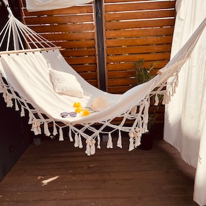 Op de afbeelding: Een witte hangmat met franjes en een houten lat hangt tussen een houten lattenwand en een wit gordijn. De hangmat is versierd met gele bloemen, een zonnebril en een strohoed.