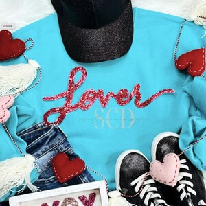 Puede incluir: Una sudadera azul turquesa con la palabra "Lover" en letras de purpurina rosa. La sudadera se combina con una gorra de béisbol negra, jeans azules y un ribete blanco peludo.