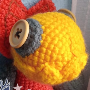 Può includere: Un giocattolo di peluche lavorato a uncinetto con uno schema di colori rosso, giallo, blu e marrone. Il giocattolo ha un bottone sulla testa e un logo di cervo sul petto. Il logo dice "Cienboazul".