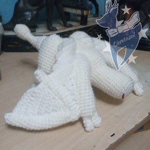 May include: White crocheted dragon plush toy with a red nose. The dragon is lying on its side with its wings spread out.