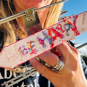 May include: A white friendship bracelet with the words "BE HAPPY" embroidered in colourful thread. The bracelet has a red border and is held in a metal frame. The background shows a sunny outdoor scene.