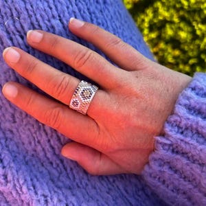 Peut inclure: Une bague en argent avec un motif de perles blanches, noires et beiges. La bague est portée sur une main, avec un pull en tricot violet en arrière-plan. Le motif de la bague comprend des motifs floraux.