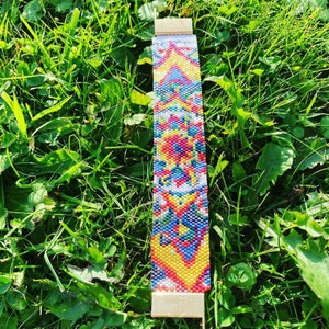 Peut inclure: Un bracelet perlé coloré avec un motif géométrique dans les tons de jaune, rouge, bleu et blanc. Le bracelet a un fermoir doré et est posé sur de l'herbe verte.
