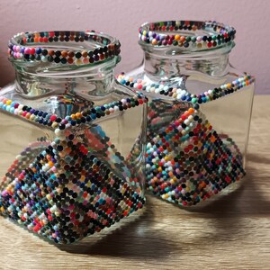 May include: Two clear glass jars with colourful beaded designs. The jars are square shaped and have a beaded pattern on the sides. The jars are empty and sitting on a wooden surface.