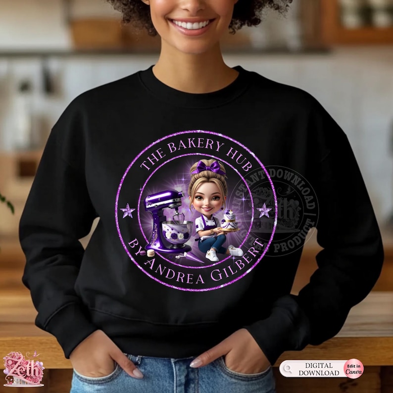 K&ouml;nnte beinhalten: Schwarzes Sweatshirt mit einem kreisf&ouml;rmigen Design. Das Design zeigt den Text "THE BAKERY HUB BY ANDREA GILBERT" und ein Cartoon-Bild einer Frau, die einen Kuchen, einen Mixer und Sterne h&auml;lt, in Lila und Wei&szlig;.