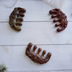 May include: Three wooden hair combs with intricate floral designs. The combs are brown, dark green, and light brown. They are arranged on a white wooden surface.