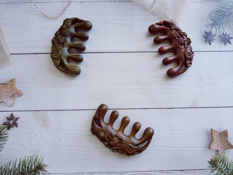 May include: Three wooden hair combs with intricate floral designs. The combs are brown, dark green, and light brown. They are arranged on a white wooden surface.