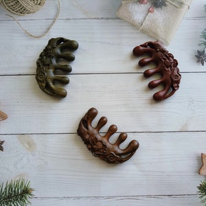 May include: Three wooden hair combs with intricate floral designs. The combs are brown, green, and dark brown in color. They are arranged on a white wooden surface.