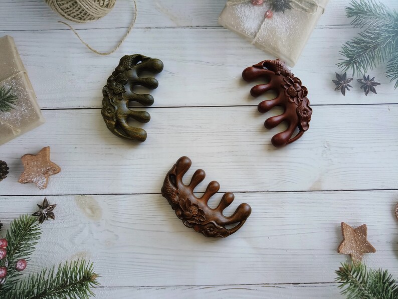 May include: Three wooden hair combs with intricate floral designs. The combs are brown, green, and dark brown in color. They are arranged on a white wooden surface.