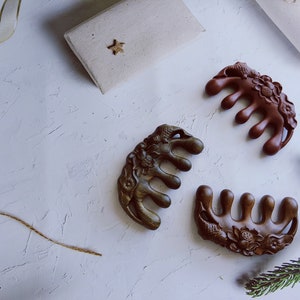 May include: Three intricately carved wooden hair combs with floral and animal designs. The combs are brown, dark brown, and reddish brown. The combs are on a white textured surface.