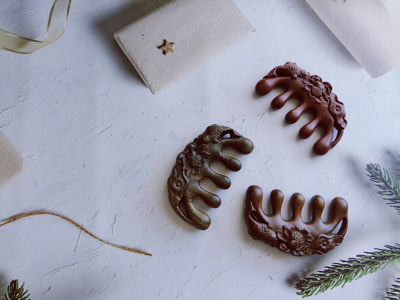 May include: Three intricately carved wooden hair combs with floral and animal designs. The combs are brown, dark brown, and reddish brown. The combs are on a white textured surface.