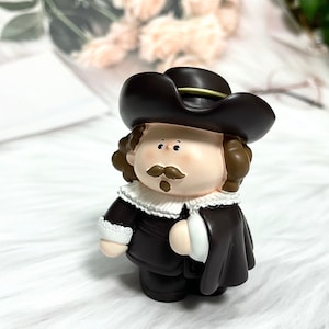 May include: A brown and white figurine of a person wearing a brown hat with a gold trim, a white collar, and a brown coat. The figurine has brown hair, a brown mustache, and a brown beard.