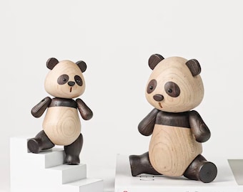 Puede incluir: Dos figuritas de panda de madera, una de pie en una escalera blanca y la otra sentada en un libro. Ambos pandas tienen detalles pintados en blanco y negro y están hechos de madera natural.