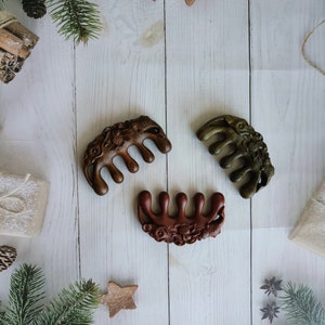 May include: Three wooden hair combs with intricate floral designs. The combs are brown, dark brown, and green. The combs are arranged on a white wooden surface with Christmas decorations.