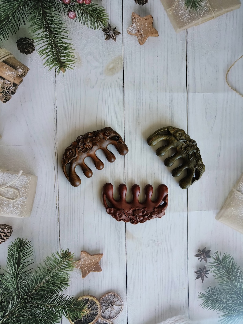May include: Three wooden hair combs with intricate floral designs. The combs are brown, dark brown, and green. The combs are arranged on a white wooden surface with Christmas decorations.
